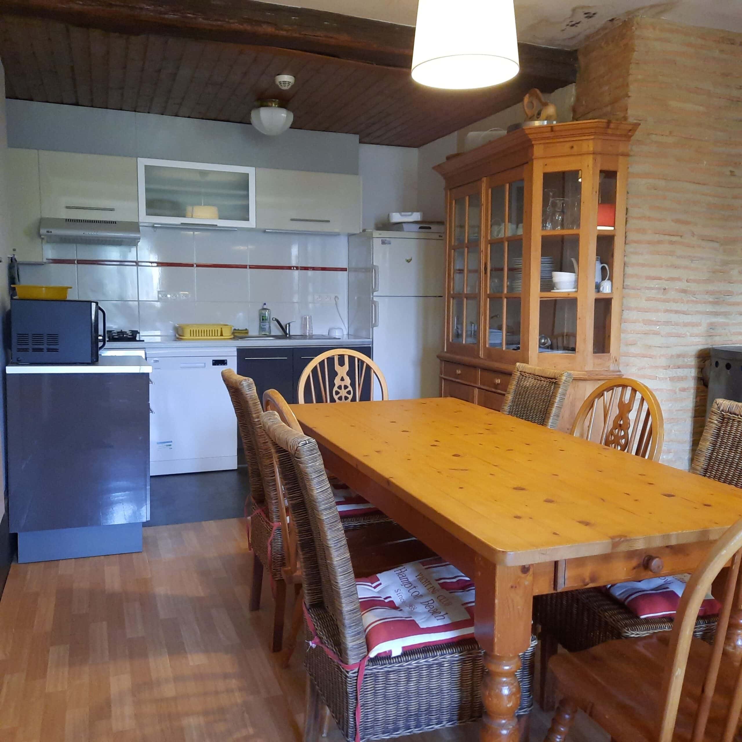 Cuisine moderne grise et blanche ouverte sur une salle à manger rustique. Grande table en bois, armoire vitrée, chaises en rotin et poutres apparentes.