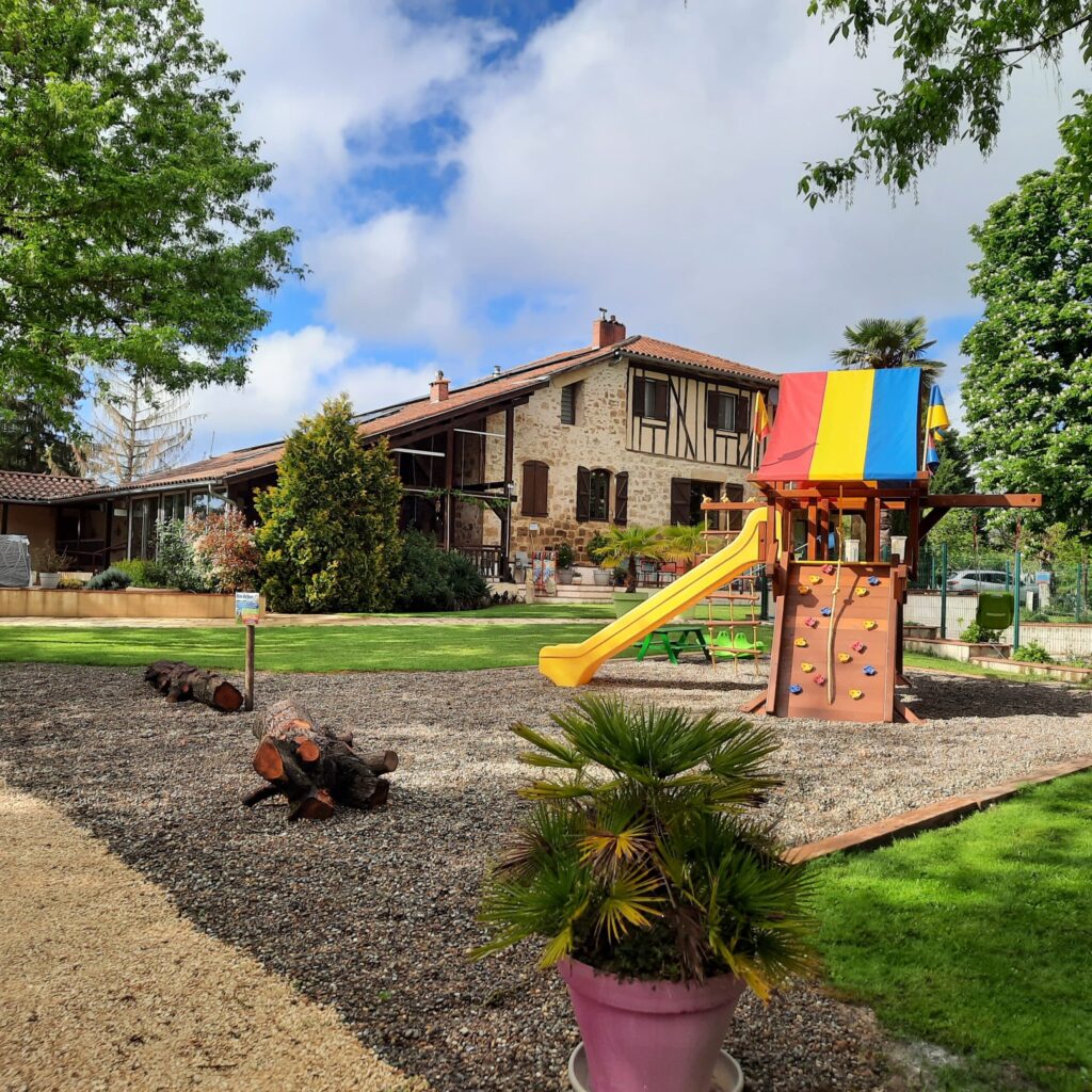 Aire de jeux enfant avec toboggan jaune devant une maison de caractère en pierre et colombages.