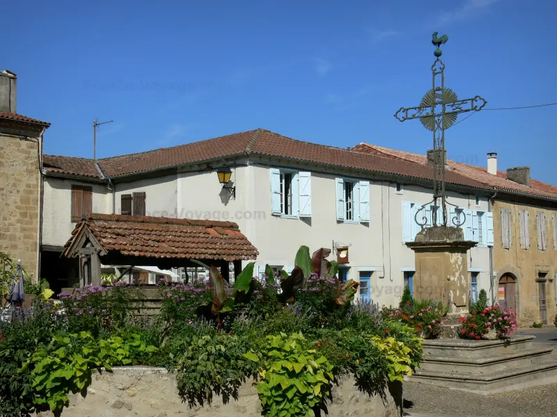 Calvaire en fer forgé, puits fleuri et maisons à volets bleus dans un charmant village français authentique.